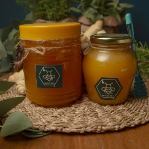 Two jars of honey on a woven mat surrounded by eucalyptus leaves and herbs.