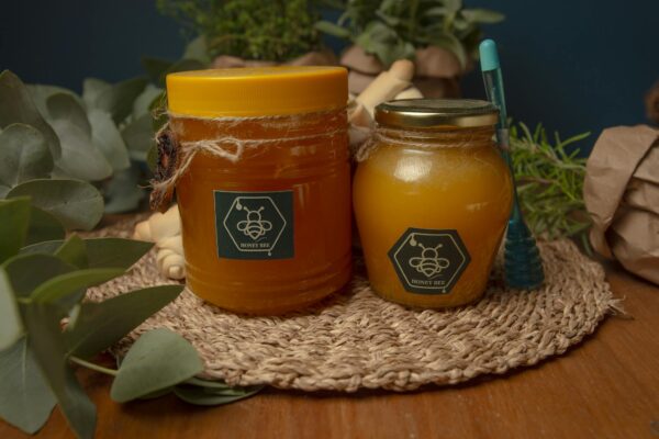 Two jars of honey on a woven mat surrounded by eucalyptus leaves and herbs.
