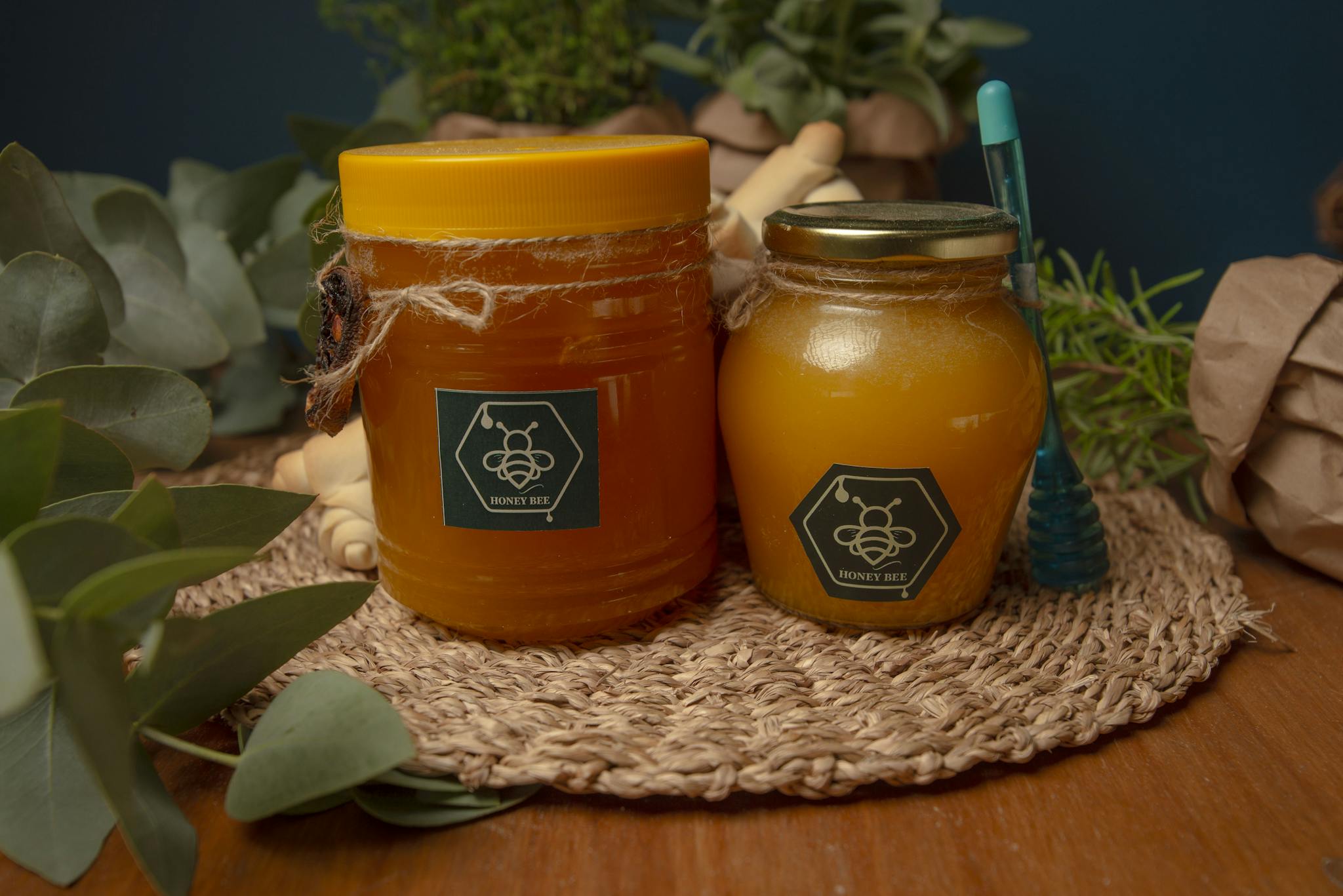 Two jars of honey on a woven mat surrounded by eucalyptus leaves and herbs.